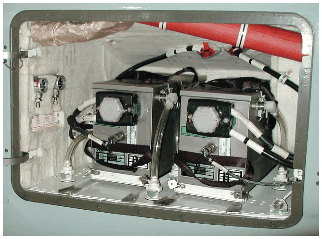 Photo of an open compartment with two boxy main batteries side by side, each with a vent cap on its front face, cables and clear vent tubes running from the cases.