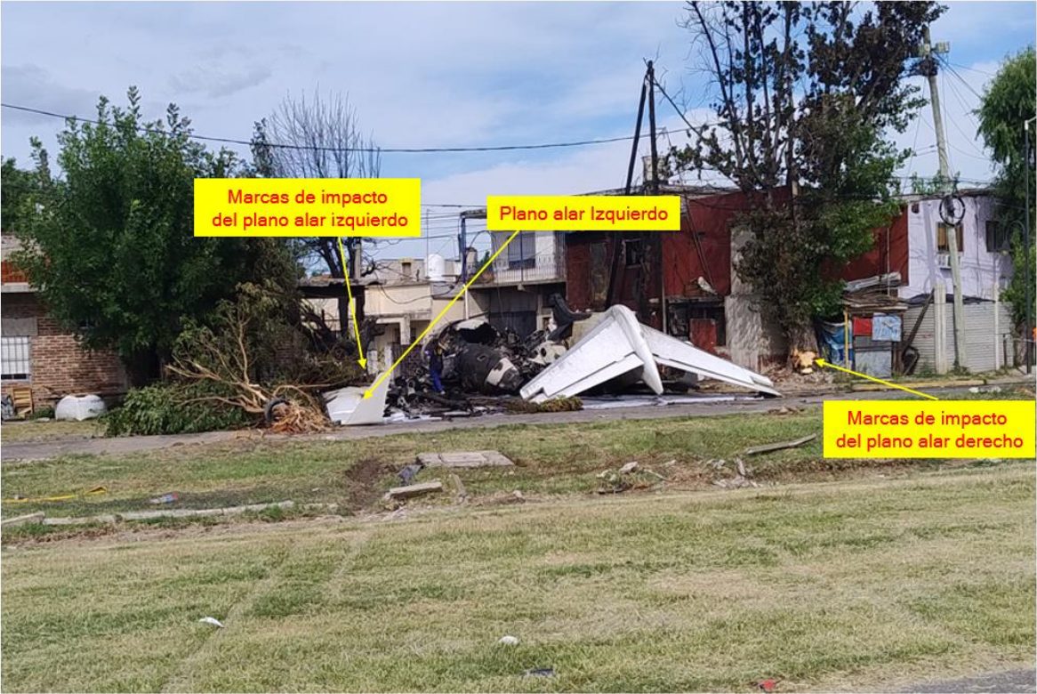 View across a grass area of the wreckage in the street, with yellow labels in Spanish marking the left wing and the impact marks of both wings on trees.